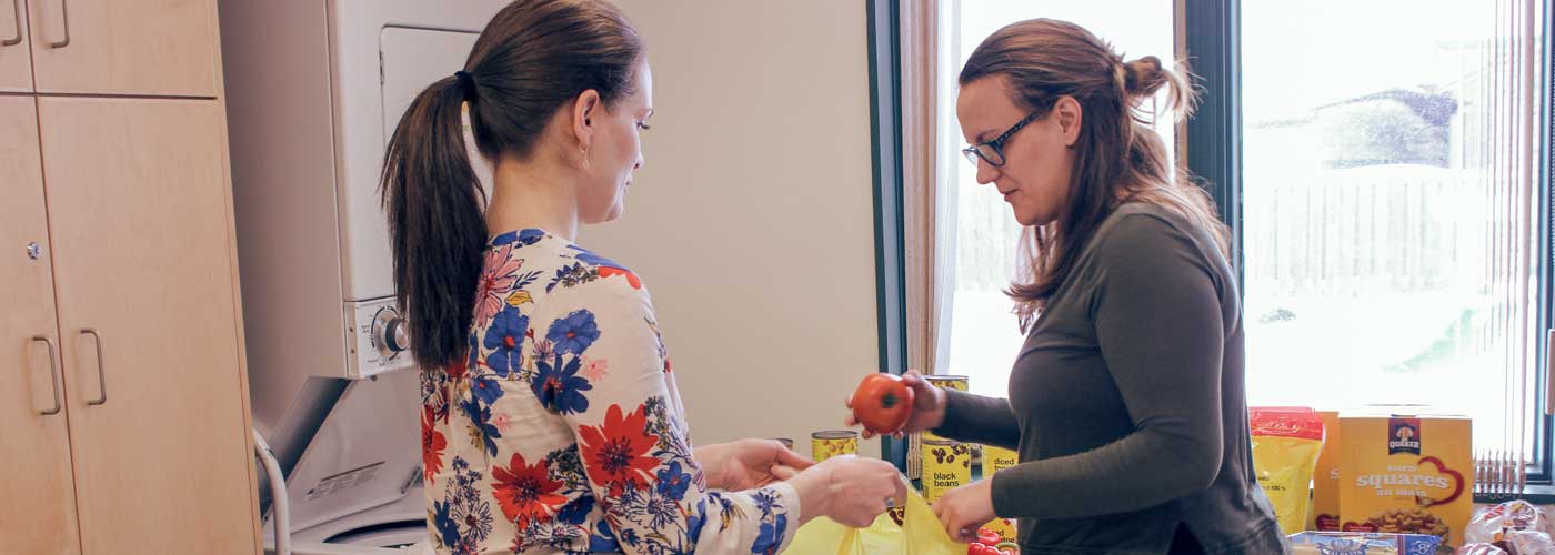 One adult holding a grocery bag, one adult putting a tomato in the bag. Canned food on a table in the background.