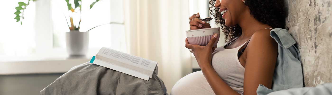 Pregnant person laying in bed reading a book and eating from a bowl