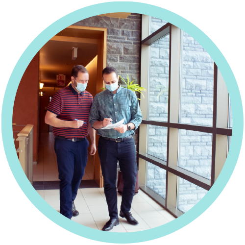 Two people walking and talking in an office hallway. Both are wearing medical procedure masks.